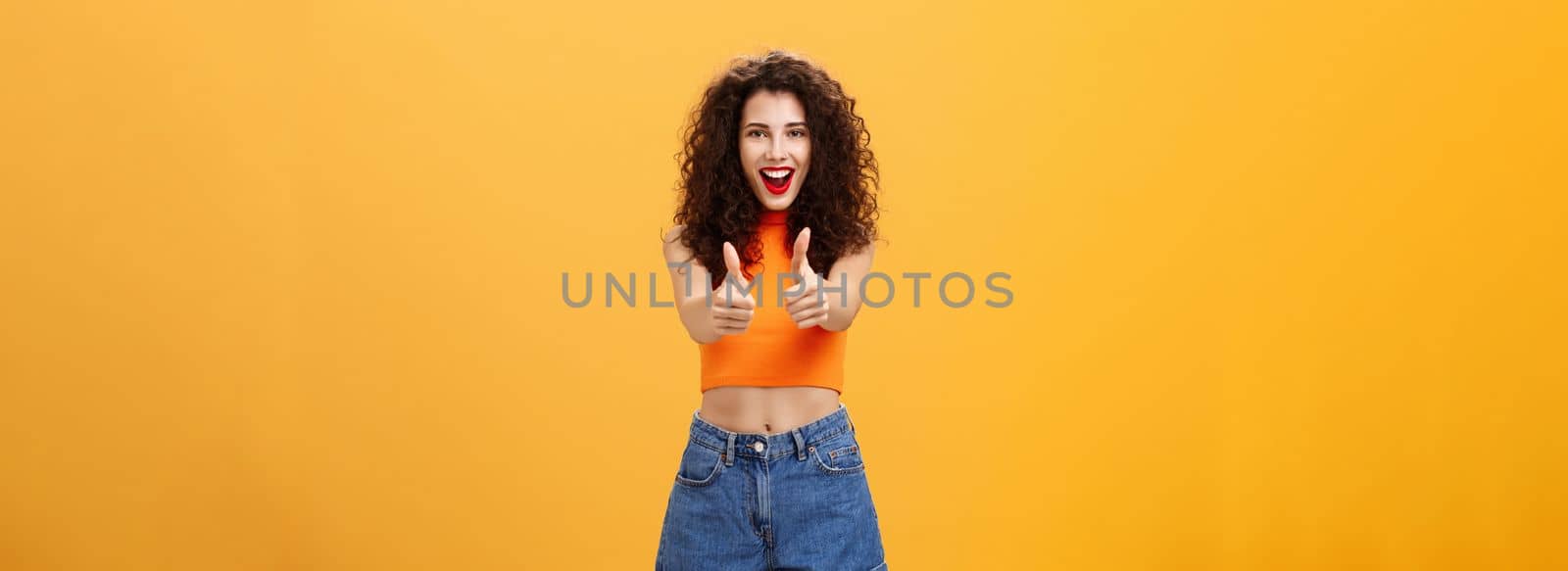 Charismatic ambitious and outgoing charming caucasian. woman with curly hairstyle and red lipstick showing thumbs up gesture in like or approval smiling joyfully being supportive over orange wall.