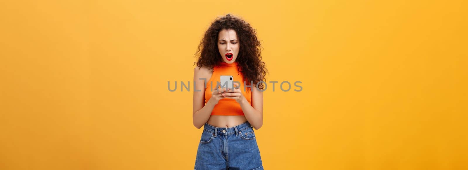 Woman being displeased and offended with low rate of photo in social network profile gasping from disappointment frowning and looking dissatisfied at smartphone screen standing over orange background. Technology concept