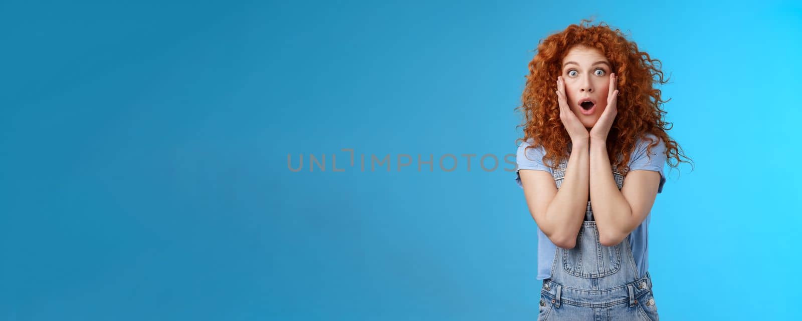 Amazed gasping shocked wondered redhead attractive curly girl popping eyes stare camera drop jaw concerned astonished touch cheeks empathy amazement standing blue background by Benzoix
