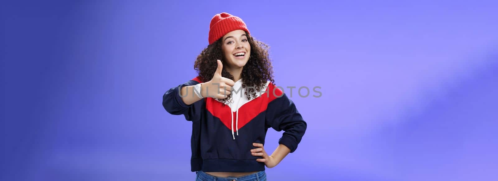 Nice work, keep up. Delighted and supportive charming sportswoman in winter beanie, sweatshirt and watch showing thumbs up with satisfied smile, giving approval, liking and cheering great effort by Benzoix