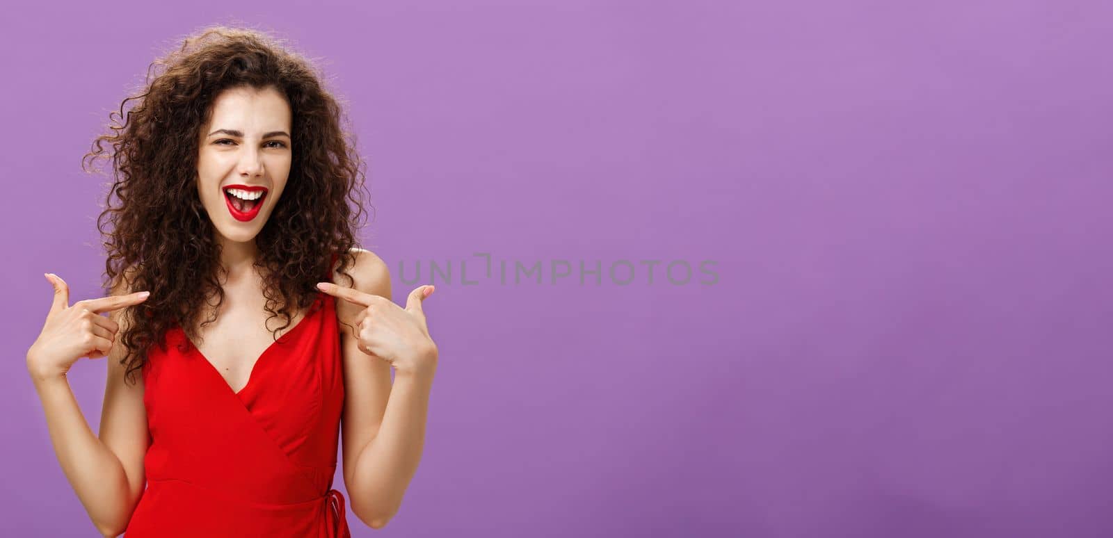 Woman celebrating successful sign of deal, pleased and satisfied bragging about own achievements pointing at herself smiling joyfully with triumph standing in evening red dress over purple background. Copy space