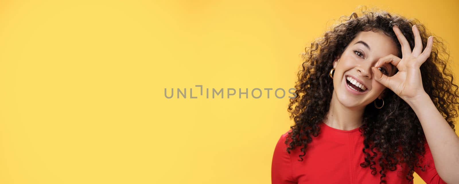 Hey I see you. Charismatic funny and playful cute tender girlfriend with curly hair showing okay or zero gesture over eye peeking at camera and smiling broadly, tilting head right over yellow wall by Benzoix