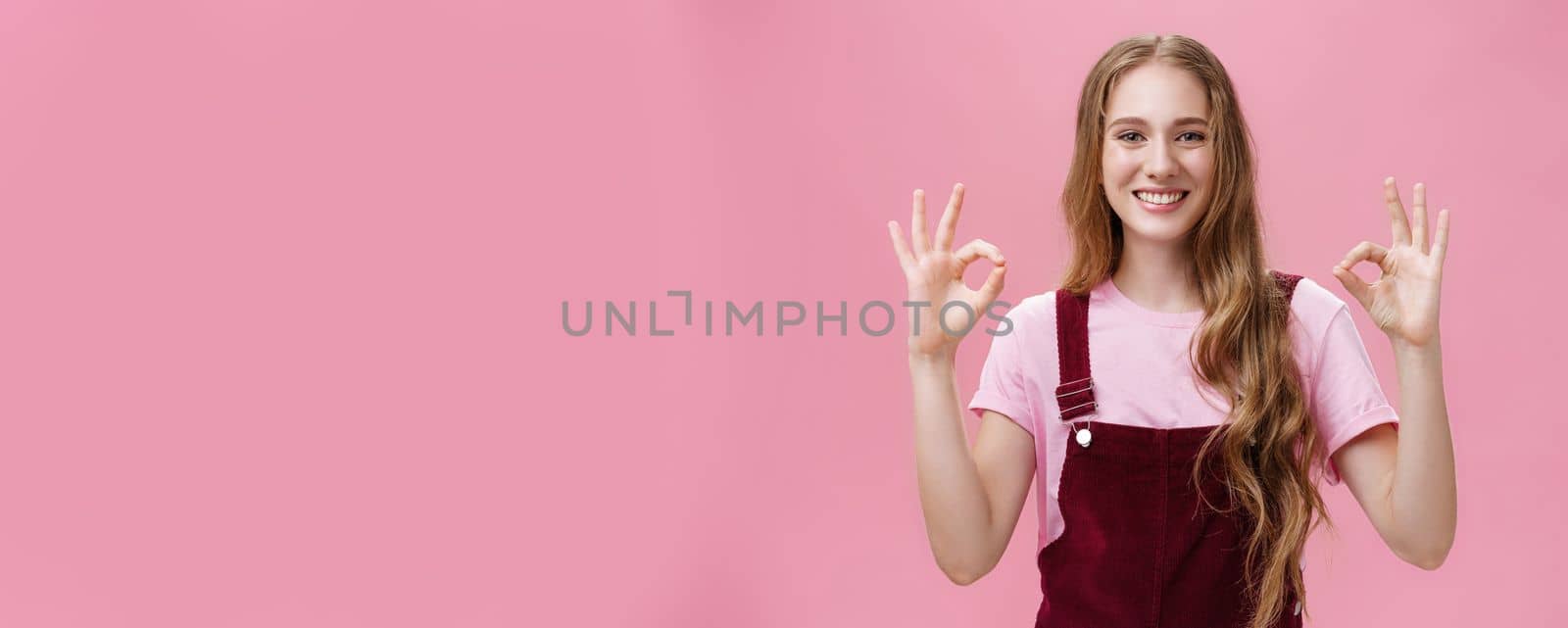 No problem, everything okay. Portrait of pleasant and kind pretty young female in dungarees showing ok or perfect gesture with both hands smiling broadly with friendly gaze at camera over pink wall by Benzoix