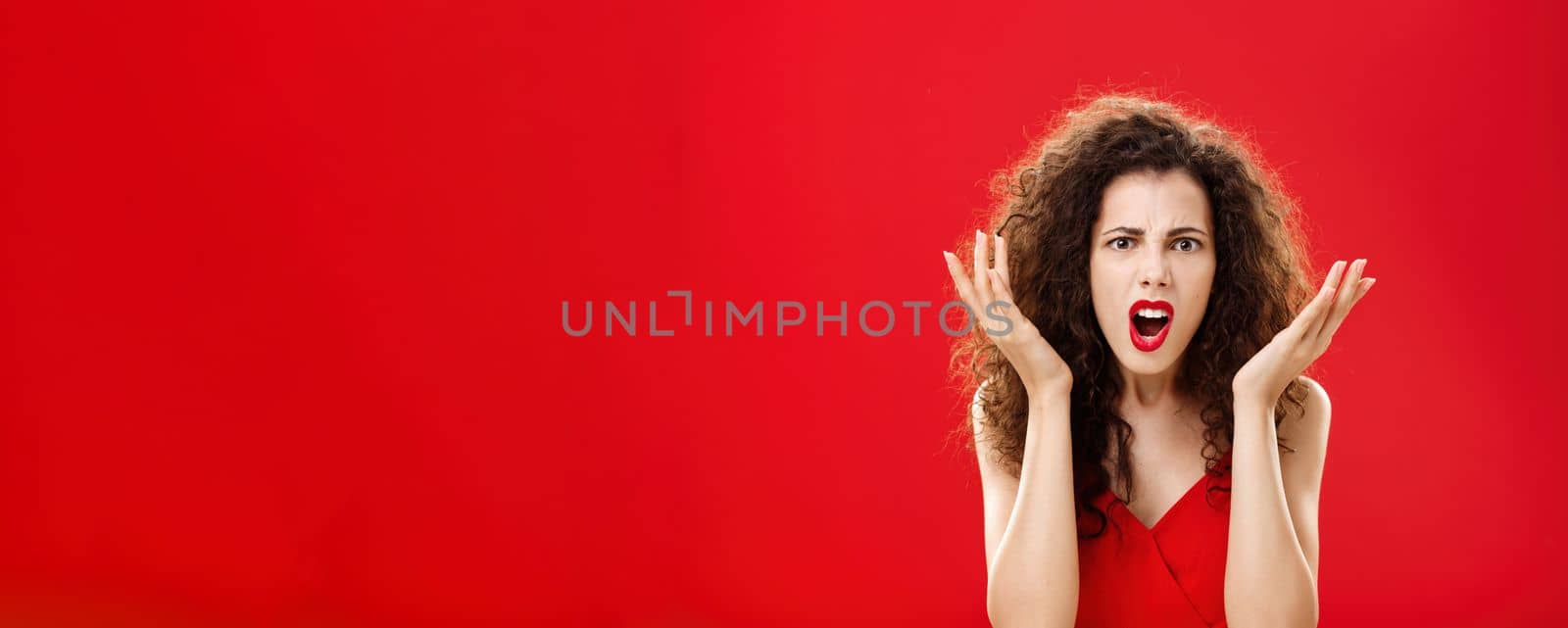 Moody displeased and angry rich woman being dissatisfied with awful chip gift raising palms in frustration and clueless gesture arguing, shouting in outrage standing in red elegant dress. Copy space