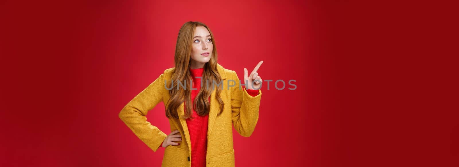 Wondered curious good-looking redhead woman in coat stopping on way looking and pointing at upper left corner curious and interested, observing surprising promotion over red background by Benzoix