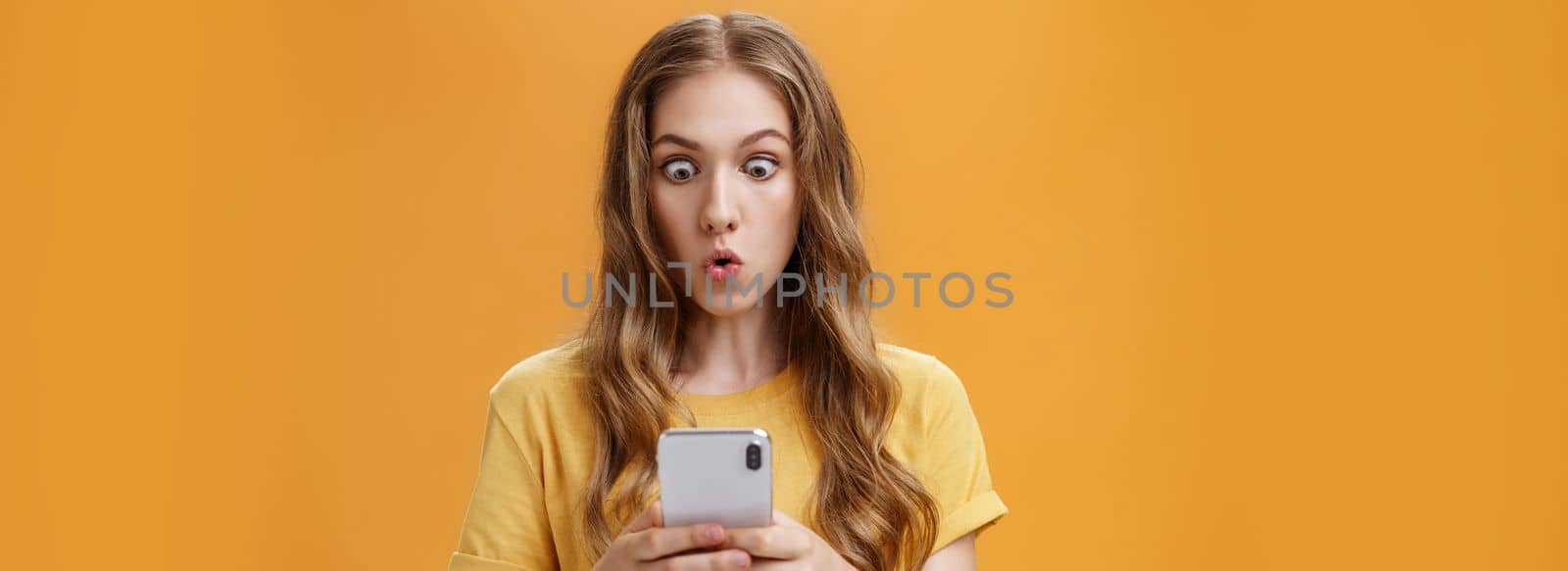 Young girl shocked reading messages in friends smartphone folding lips popping eyes in amazement at cellphone screen folding lips with interest and excitement posing over orange background. Emotions and technology concept