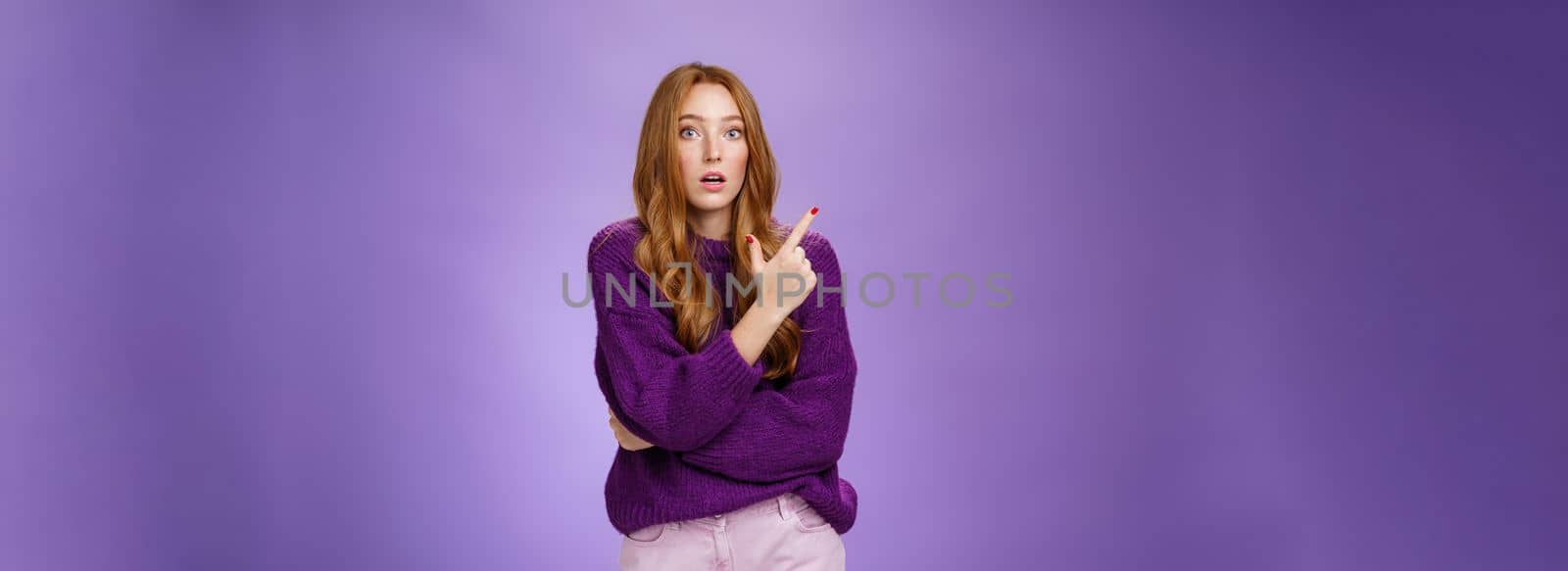 Astonished excited redhead woman open mouth and raise eyebrows questioned as seing shocking thing pointing at upper left corner, asking question impressed and surprised over purple wall.