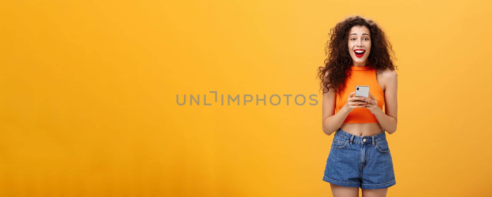 Indoor shot of surprised happy and delighted attractive european female with curly haircut in cropped top holding smartphone gazing at camera amazed and pleased receiving awesome news via messages. Technology concept