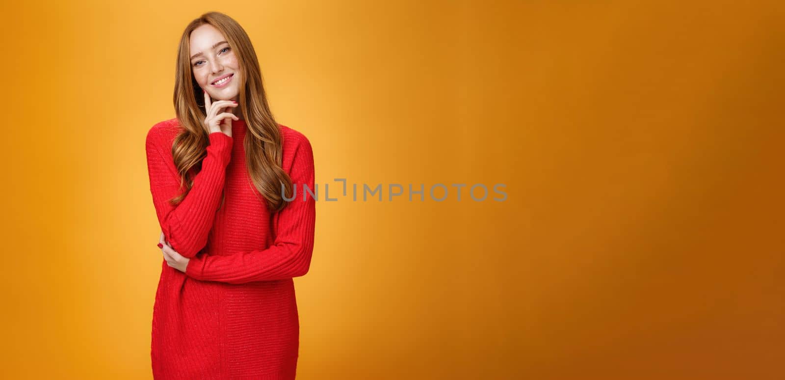 Sweet and tender redhead woman with cute freckles in red knitted dress tilting head touching face with fingers and smiling delighted and loving a camera over orange background. Copy space