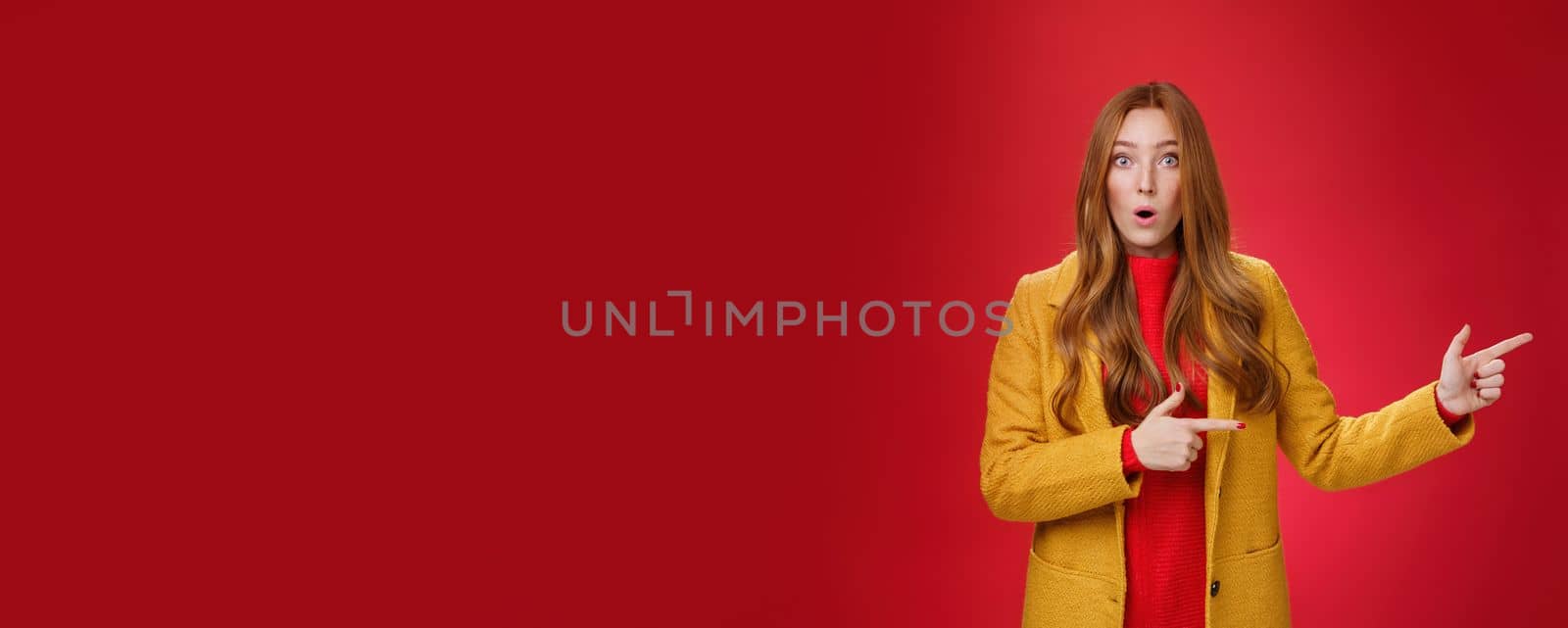 Speechless questioned and confused surprised redhead girl in yellow autumn coat pointing right open mouth curiously and staring at camera puzzled and clueless waiting explanation over red wall by Benzoix