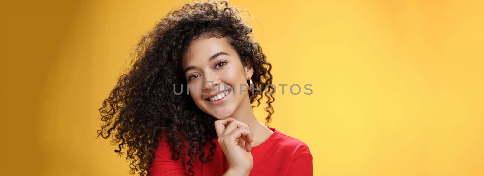 Close-up shot of stylish and happy bright curly-haired female in red dress tilting head sensually touching chin with finger and smiling broadly making flirty gazed at camera over yellow background by Benzoix