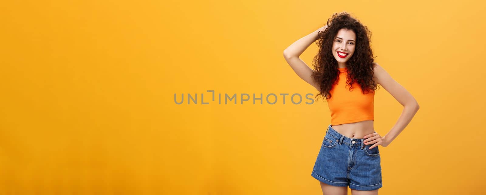 Friendly and stylish modern curly-haired female. with red lisptick in denim shorts and cropped top playing with hair smiling flirty and carefree holding hand on waist posing over orange background.