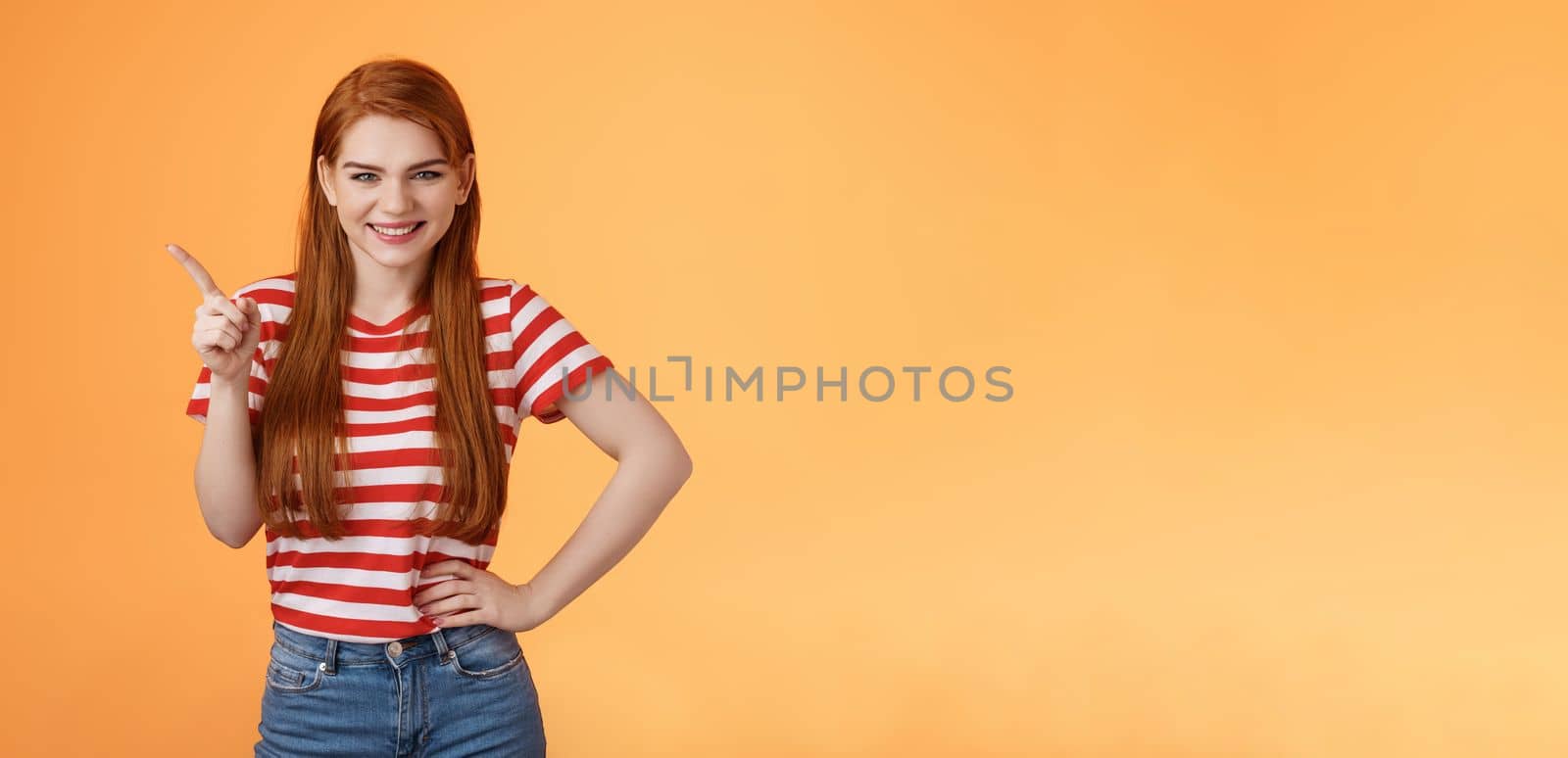 Cheerful sly redhead girlfriend have something interesting on mind, squinting cunning joyful smile, hold hand waist confident pose, pointing upper left corner give recommendation, orange background.