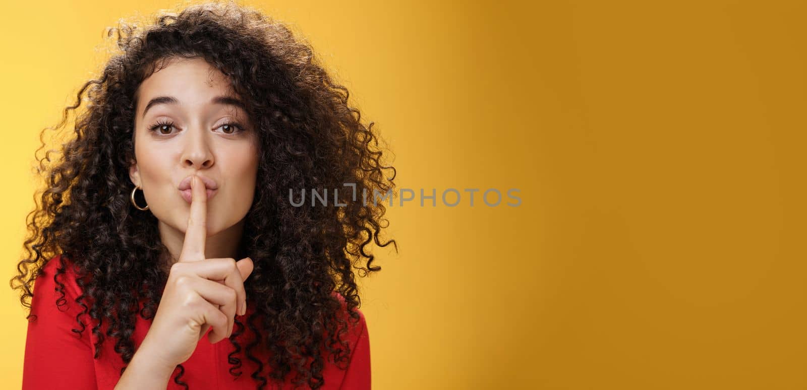 Shh please not tell secret. Cute and tender woman with curly hairstyle gently shushing with index finger on folded lips asking keep promise and remain silent, making surprise over yellow wall by Benzoix