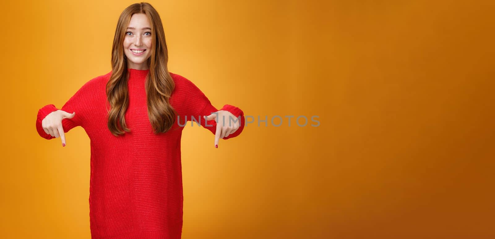 Gentle and tender cute ginger girl with freckles and blue eyes feeling touched and pleased, pointing at heartwarming promotion, showing direction down and smiling against orange background.
