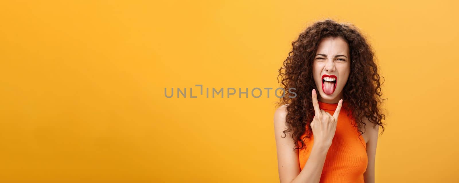 Carefree rebellious stylish urban female with curly hairstyle sticking out tongue yelling from satisfaction and joy showing rock n roll sign chilling and enjoying cool concert over orange background by Benzoix