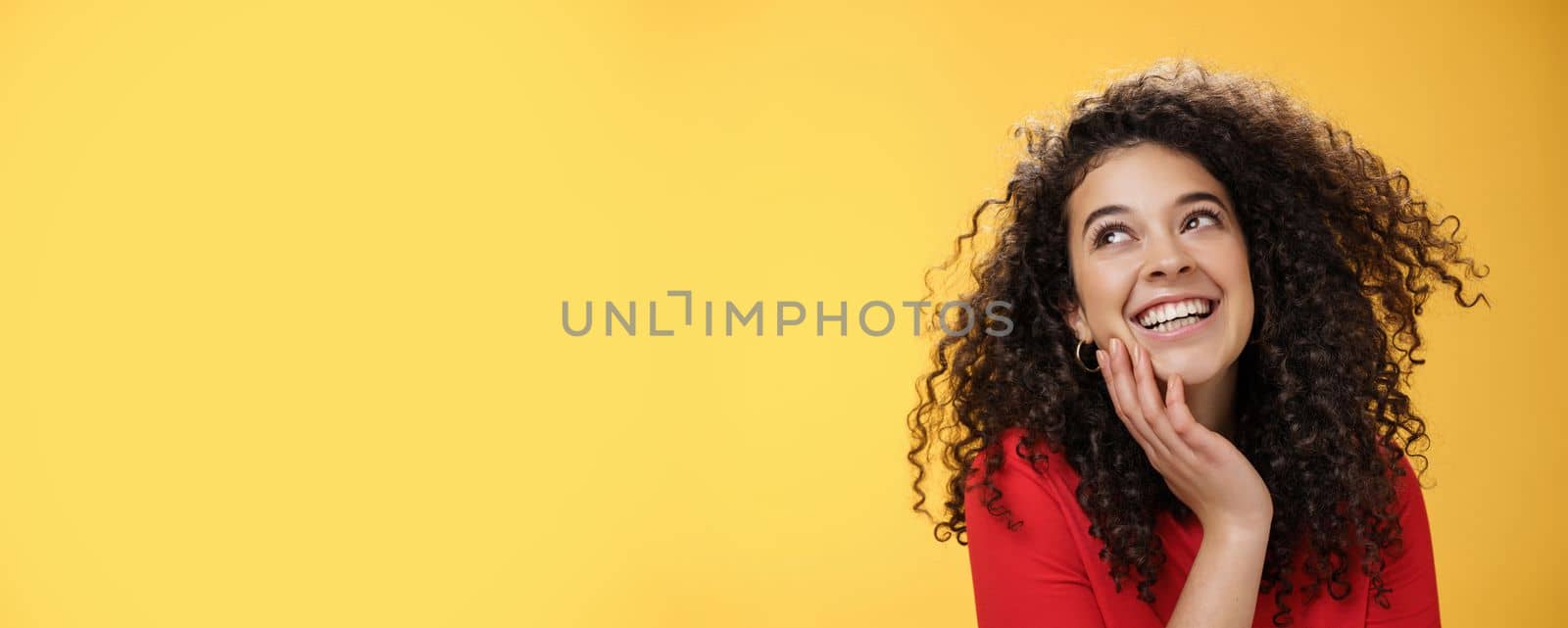 Happy positive pretty young woman with curly hair in red blouse laughing silly and carefree as gazing pleased at upper left corner, touching face, satisfied and delighted over yellow wall.