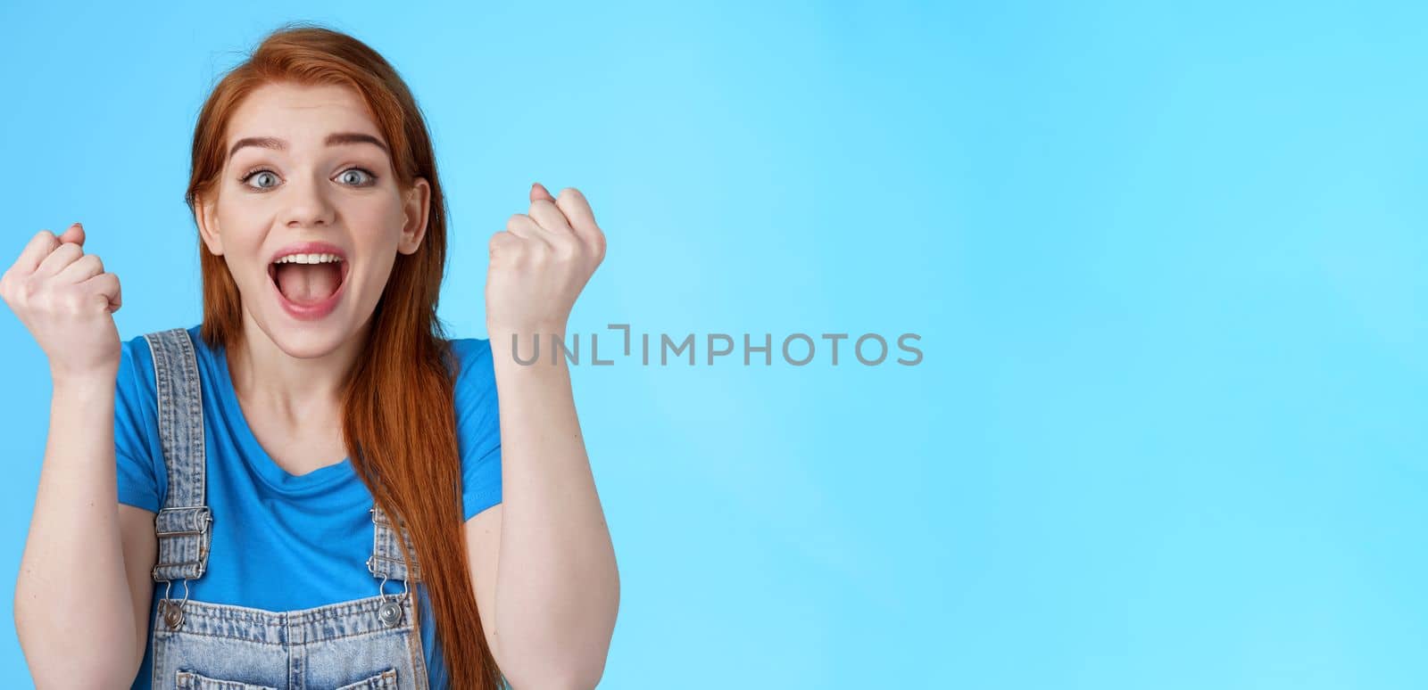 Close-up cheerful lucky redhead woman win lottery, fist pump yell yeah hooray, winning bet, smiling gladly rejoicing excellent news, celebrating success, triumphing happily, stand blue background.