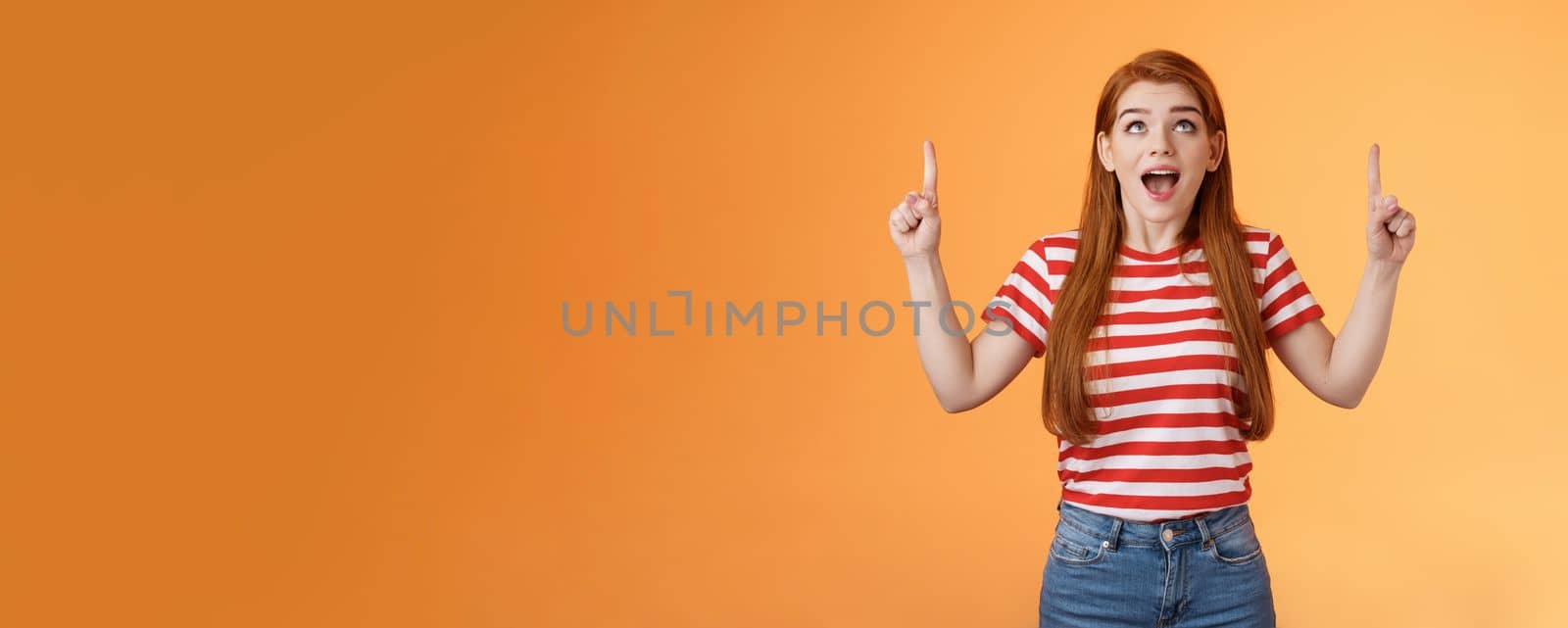Surprised excited cheerful ginger girl see amazing opportunity, look pointing up amused, drop jaw gasping astonished, pleased awesome cool advertisement, reacting thrilled top copy space.