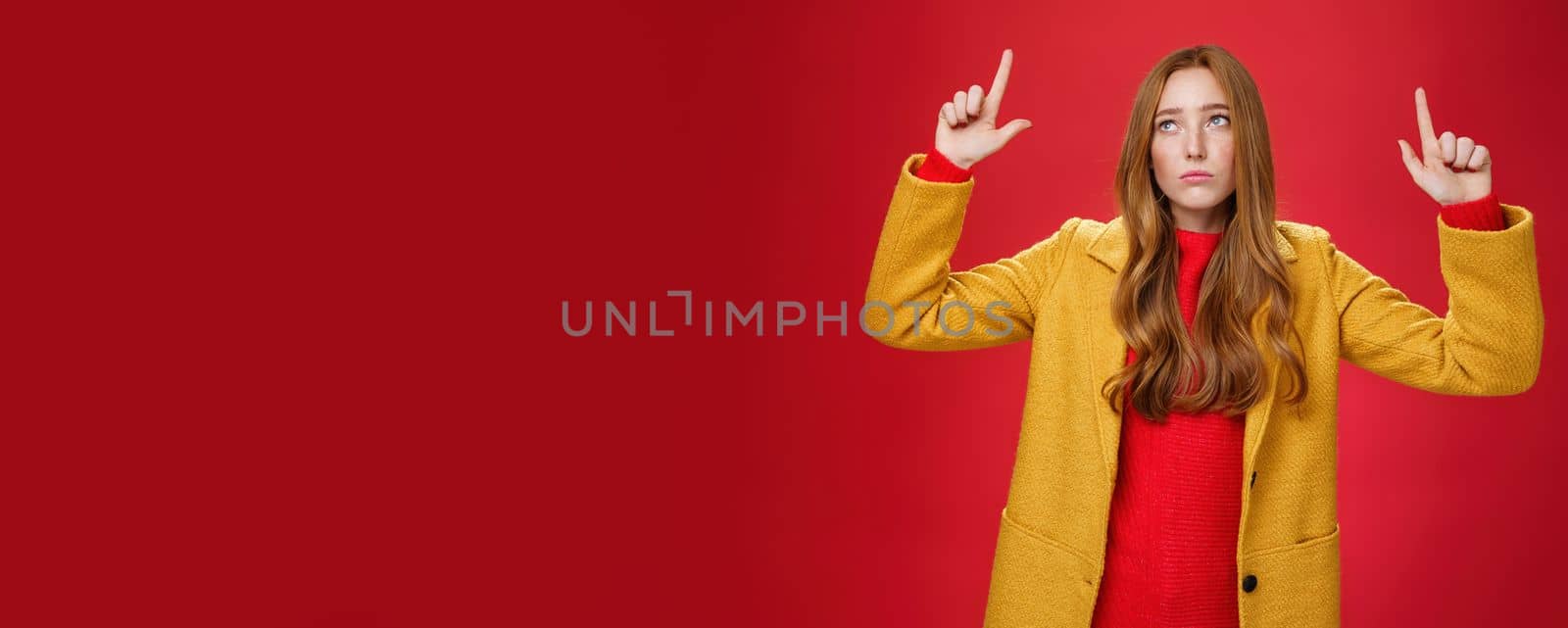 Lifestyle. Sad and sorrow cute redhead female in yellow coat raising hands up displeased with regret looking at upper left corner apathical as being disappointed with rain or bad weather over red wall.