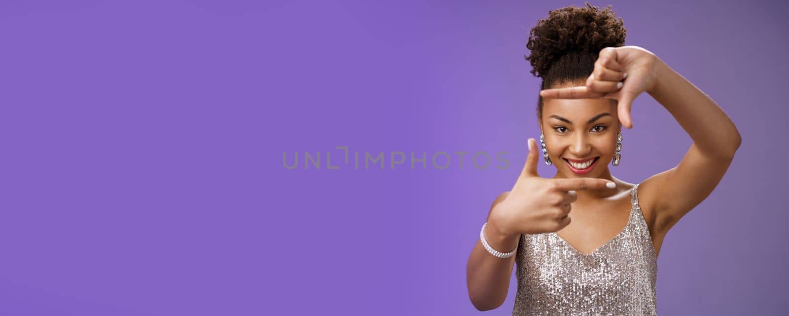 Creative good-looking african woman in silver glittering dress searching inspiration make finger frame look camera pleased smiling satisfied found perfect photo angle, standing blue background.