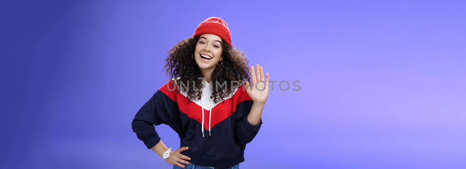 Hey nice to meet you. Charming female ski couch in cute red beanie with curly hair waving hello with palm and smiling broadly greeting newbies as teaching winter sports over blue background by Benzoix