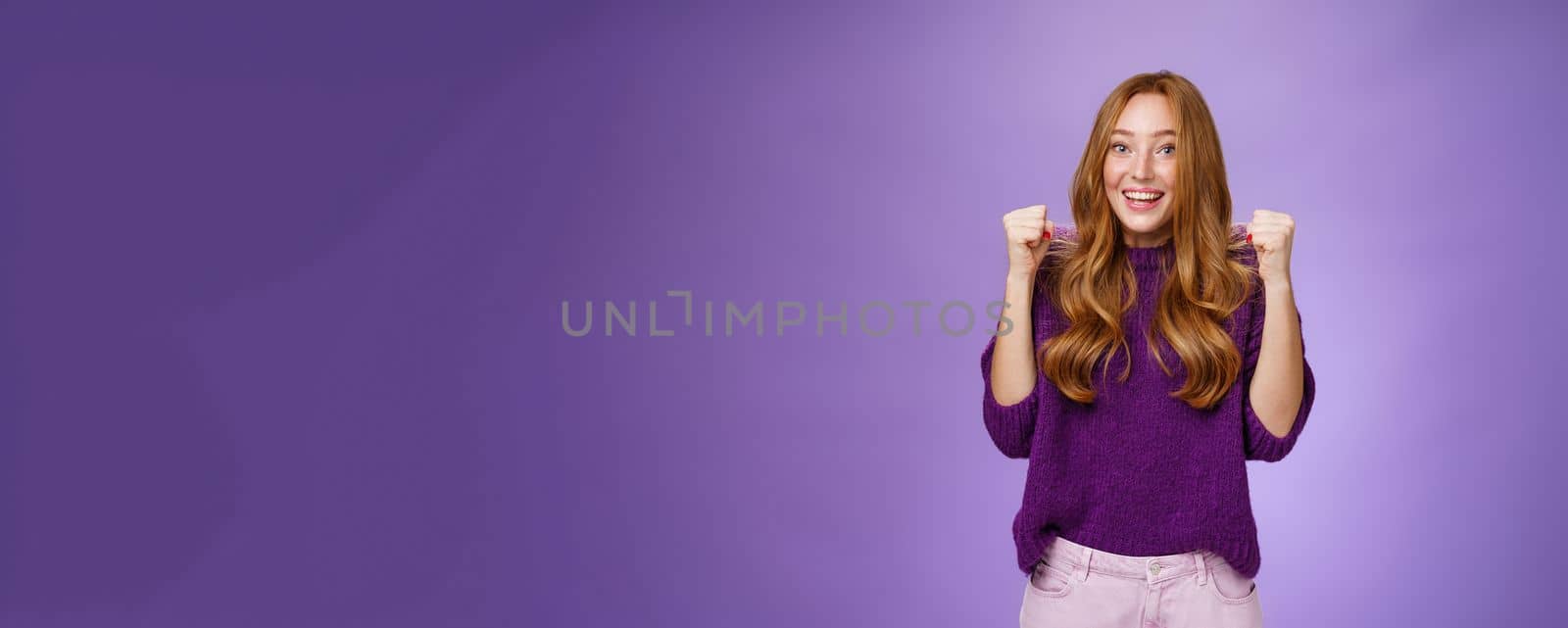 Yey we made it. Happy and cheerful young pretty redhead 20s girl in purple sweater over violet background raising clenched fists in joy and triumph, celebrating success and win, cheering. Body language and emotions concept