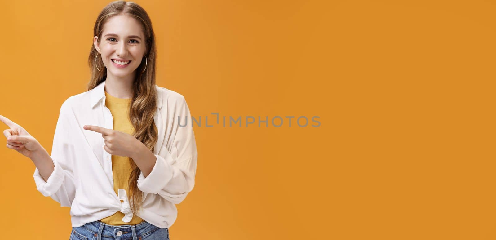 Lifestyle. Waist-up shot of friendly pleasant and stylish young teenage female in blouse over t-shirt with cute wavy fair hair pointing left and smiling broadly at camera posing happy against orange background.