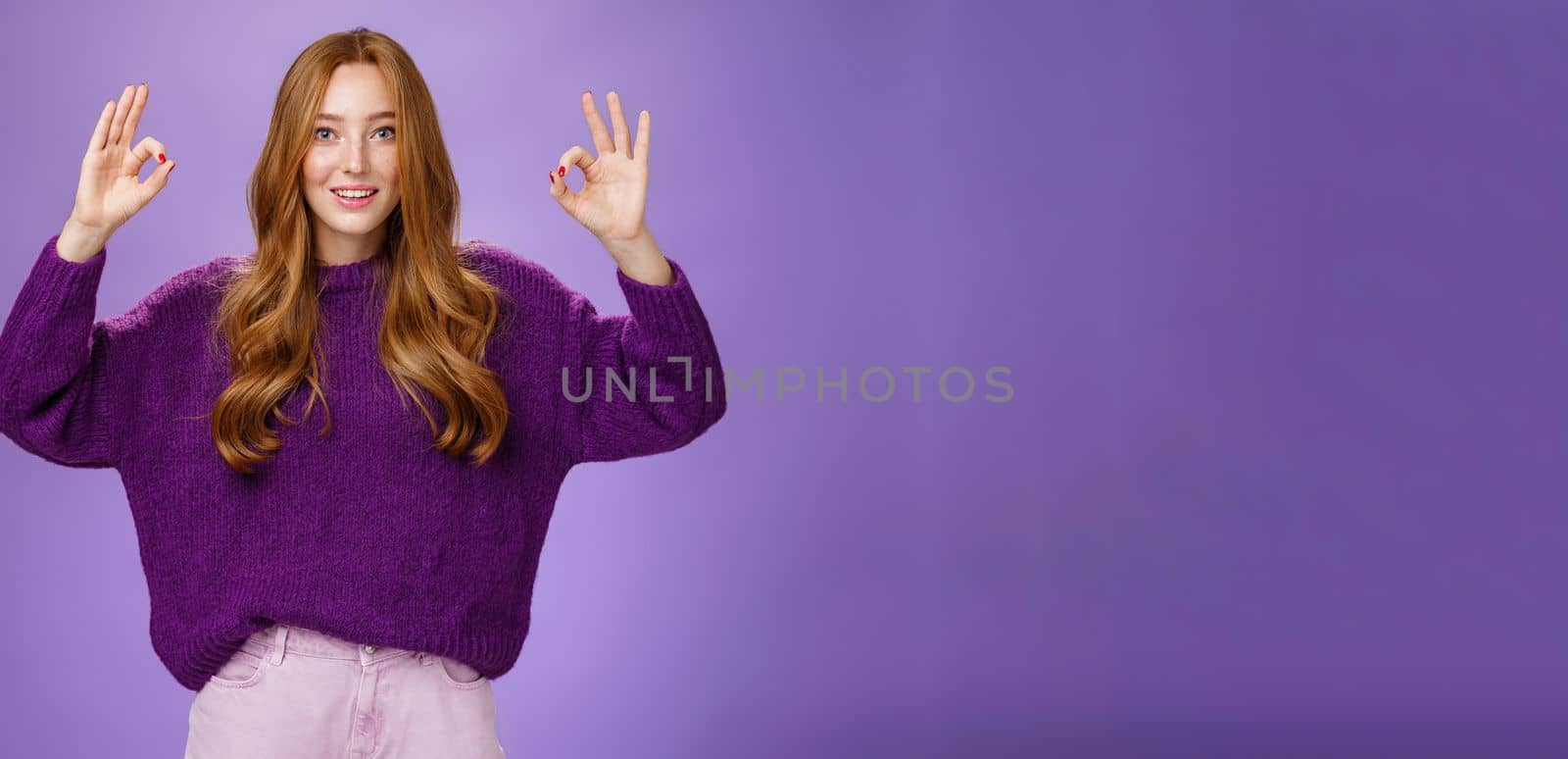 Fine I agree. Portrait of friendly and optimstic young 20s ginger girl in purple sweater raising hands with okay or ok gesture smiling in approval, liking cool product, giving recommendation.