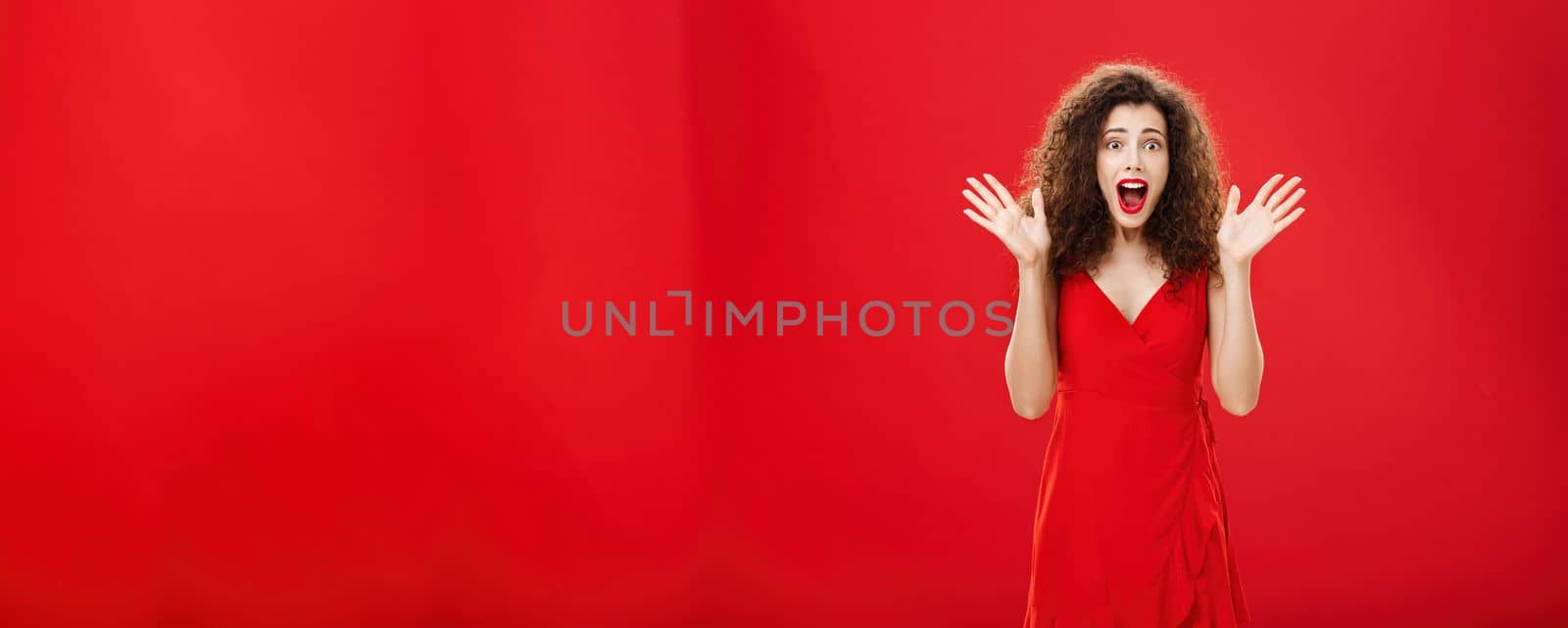 Passionate amazed and thrilled european adult woman in evening dress with curly hairstyle screaming from delight and surprise waving with raised hands and opening mouth from amazement over red wall. Copy space