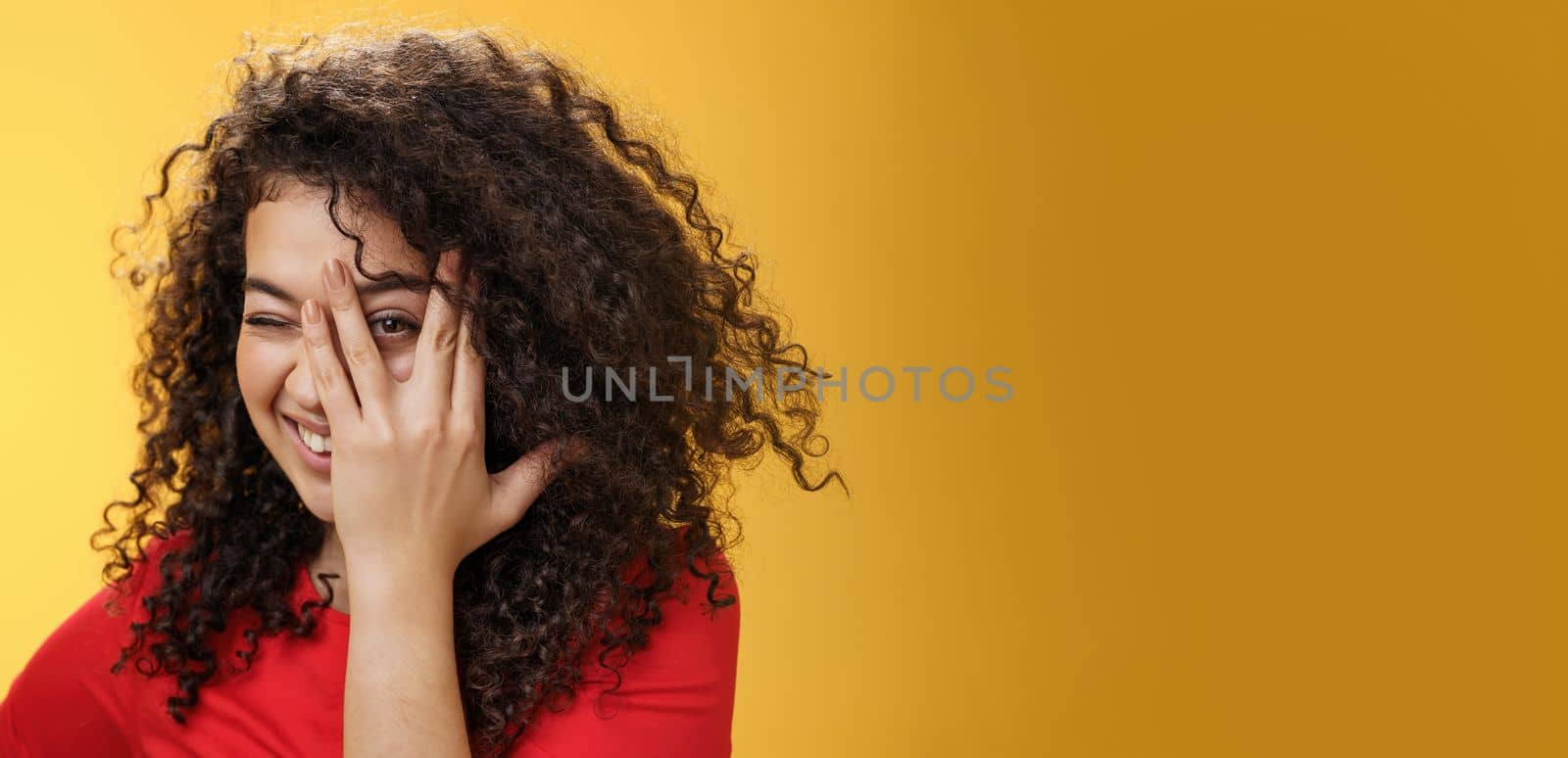 Lifestyle. Close-up shot of sensual and playful attractive girlfriend with curly hair covering face with palm and peeking through fingers with happy tender smile anticipating surprise over yellow background.