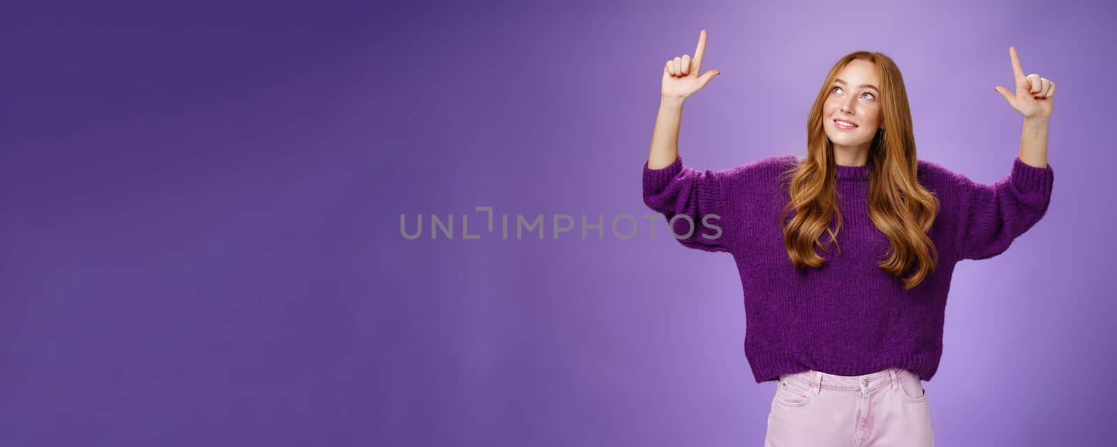 Dreamy girl with red hair and freckles in warm cozy purple sweater raising hands looking and pointing up with intrigued and happy charmed expression smiling as liking curious product over violet wall by Benzoix