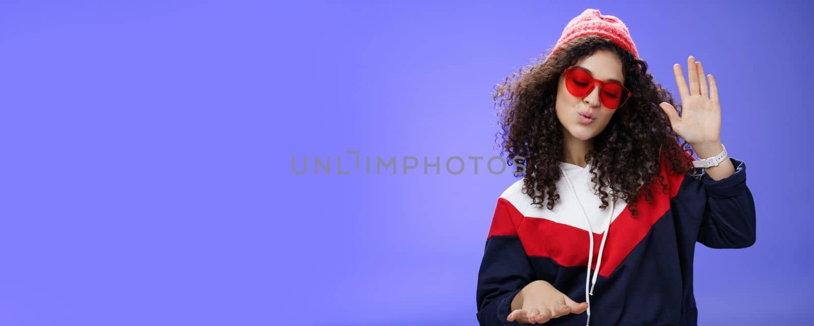 Lifestyle. Studio shot of cool and stylish dj girl in red beanie and sunglasses raising palm as enjoying cool music and dancing to rhythm folding lips having fun at awesome party posing chill over blue wall.