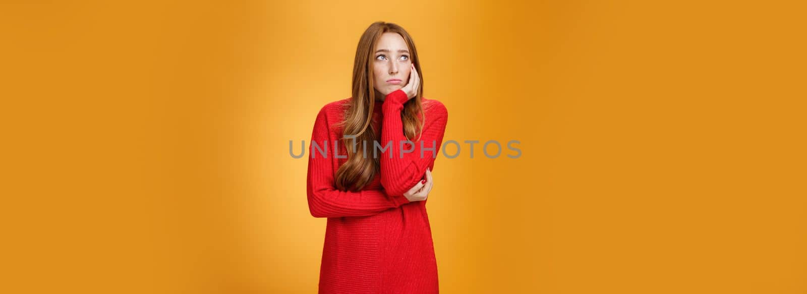 Lifestyle. Sorrow and upset lonely woman leaning head on palm in upset pose looking gloomy at upper left corner as remembering unhappy story or being bored standing uneasy over orange wall.