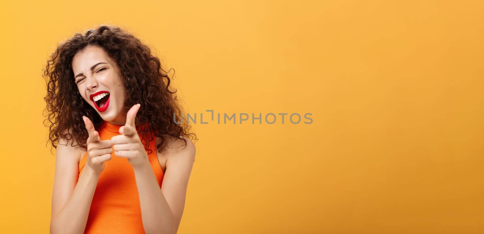 Hey what is up. Friendly and outgoing attractive curly-haired female with red lipstick pointing at camera with finger guns as if greeting or hinting friend winking at camera over orange background by Benzoix