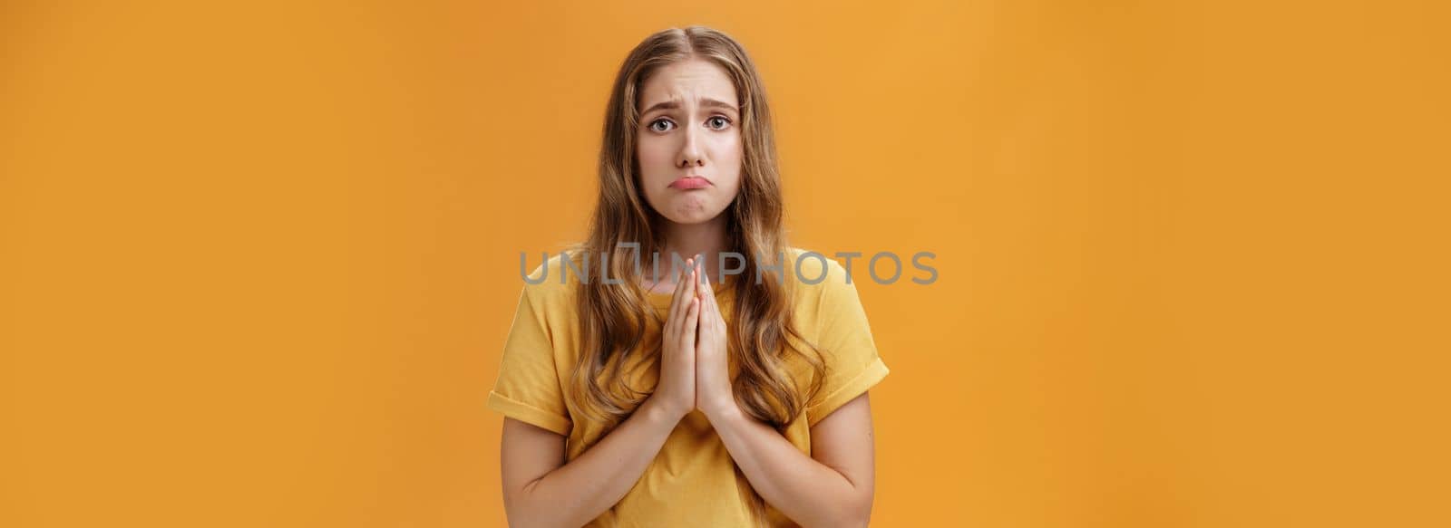 Silly girl needs help begging with hopeful puppy eyes holding hands in pray pouting and frowning feeling miserable stuck in bad situation standing upset over orange background, borrowing money.