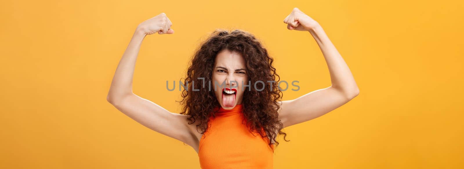 Waist-up shot of cool and daring caucasian female in orange top with tattoo on arm frowning making funny face sticking out tongue raising hands showing muscles feeling power and strengths. Copy space