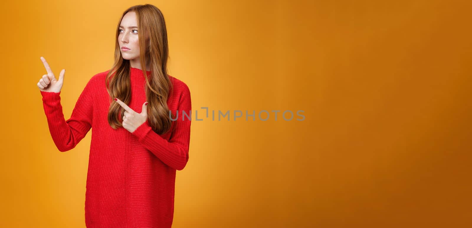 Studio shot of suspicious serious-looking unsure redhead girl frowning looking and pointing at upper left corner with doubtful strict gaze posing uncertain over orange background. Copy space
