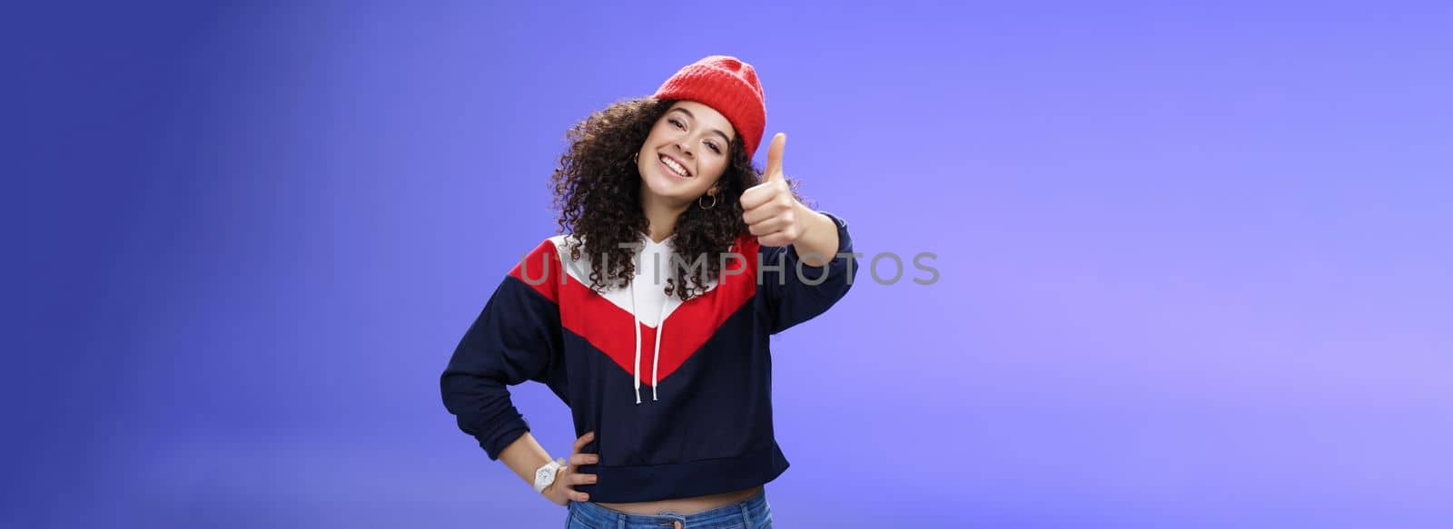 You doing great, like. Portrait of happy delighted young stylish woman in winter beanie and sweatshirt showing thumbs up pleased and satisfied approving and being supportive smiling at camera by Benzoix