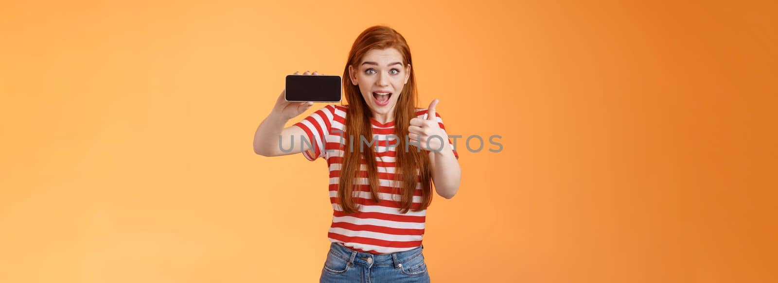 Excited cheerful 20s caucasian ginger girl, satisfied awesome game, beat friend score arcade, show thumb-up, smiling thrilled, enthusiastic introduce cellphone app, hold device horizontal.