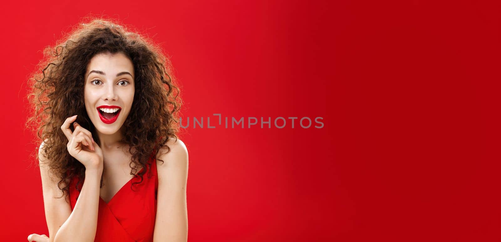 Impressed and excited feminine adult woman with curly hair in red lipstic and elegant evening dress gasping and smiling with surprised, amazed look playing with hair strand listening carefully story. Emotions concept