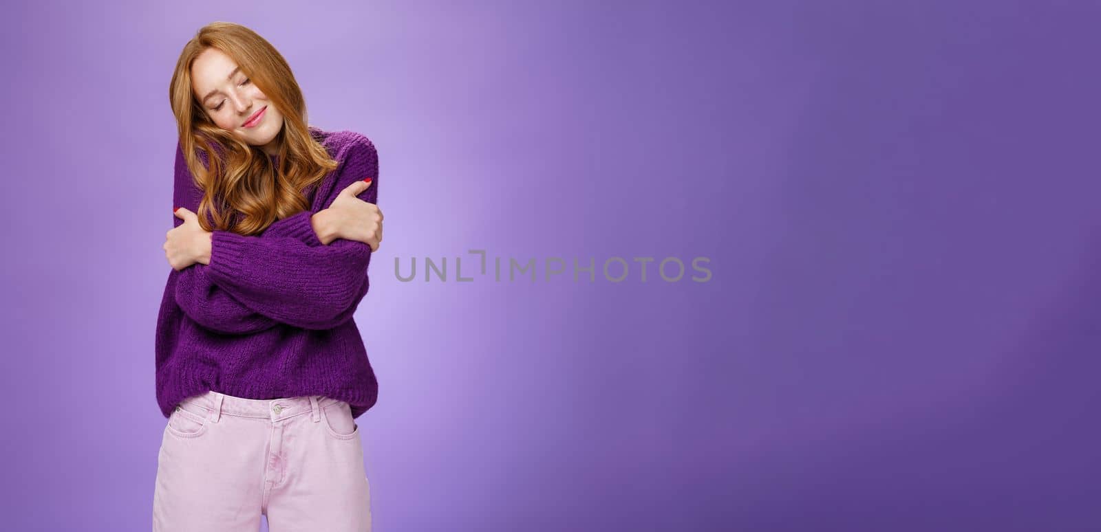 Lifestyle. Sweet and tender charming redhead girl with freckles leaning on shoulder as cross arms over chest and hugging herself liking soft and comfortable warm sweater close eyes dreamy over purple wall.