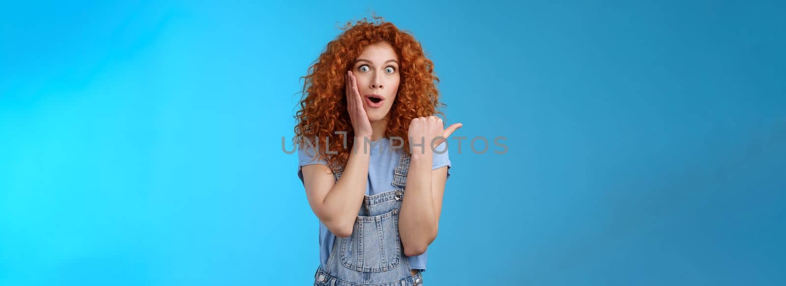 Omg how intriguing. Impressed charismatic emotive surprised redhead cute curly girl open mouth gasping curious amazed touch cheek silly pointing left thumb telling about astonishing store.