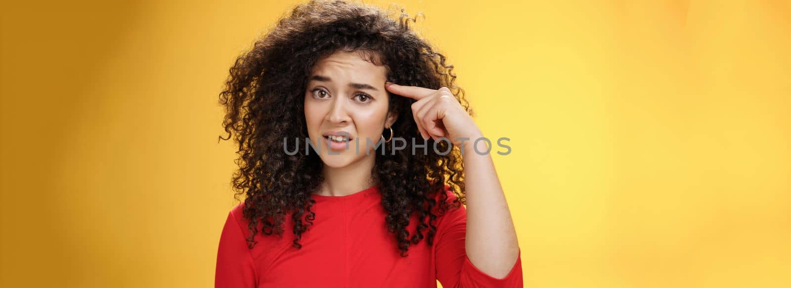 Are you crazy. Portrait of frustrated and confused woman with curly hair holding index finger near temple and raising eyebrows shocked being annoyed with stupid actions of weirdo over yellow wall.