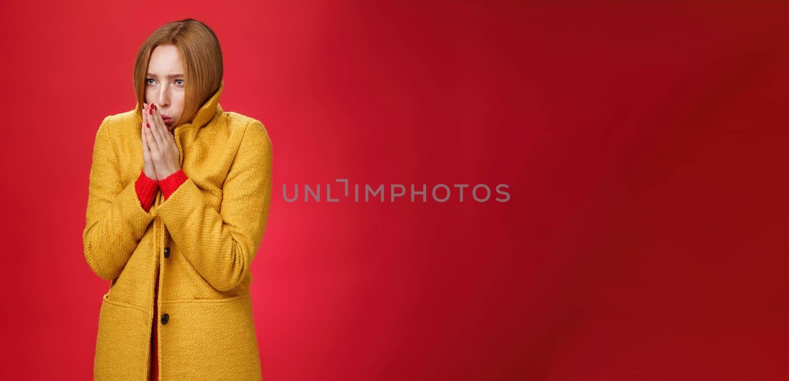 Studio shot of intense trembling cute redhead woman wearing yellow light coat rub hands and blowing warm breath at palms to warm-up feeling cold, freezing during cold windy weather over red background.
