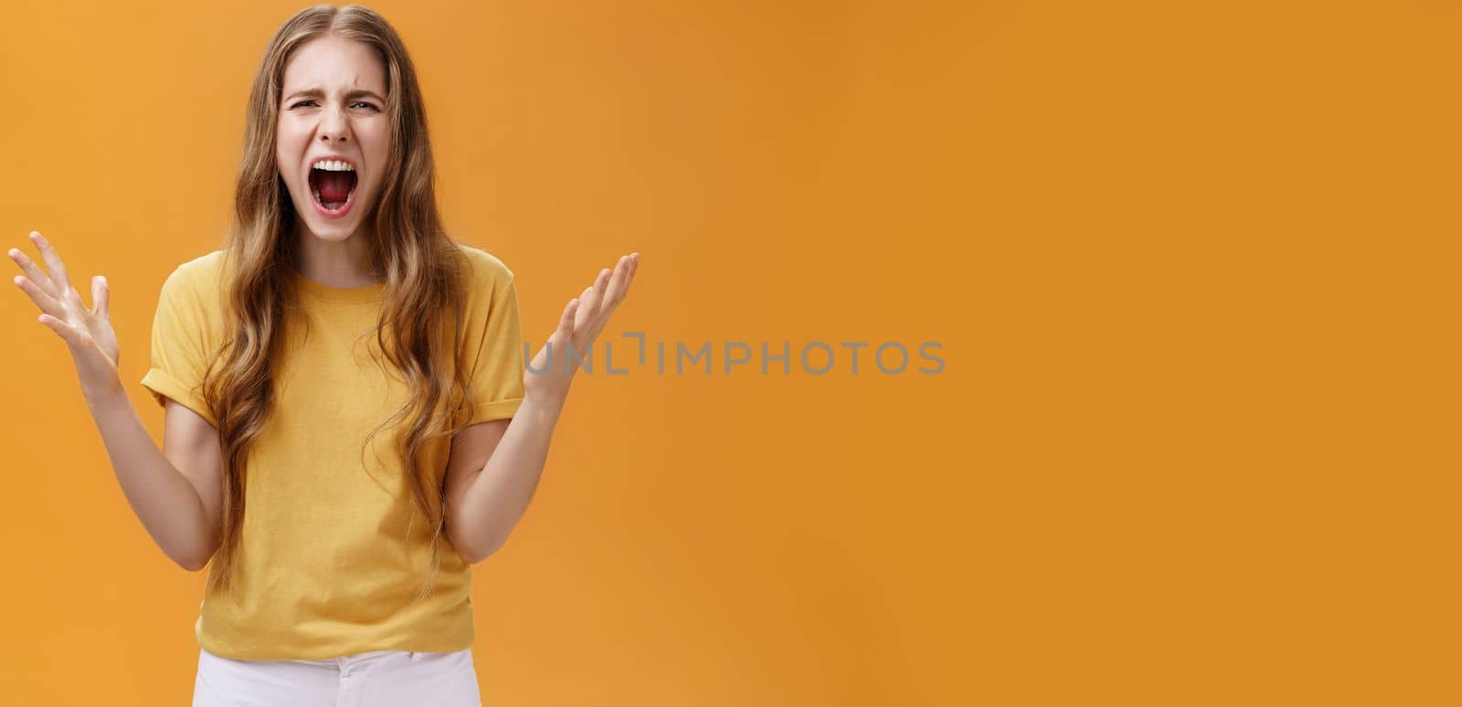 Studio shot of young woman during argument losing temper standing pressured and pissed yelling out loud from hate and anger gesturing with raised palms making furious face posing against orange wall by Benzoix