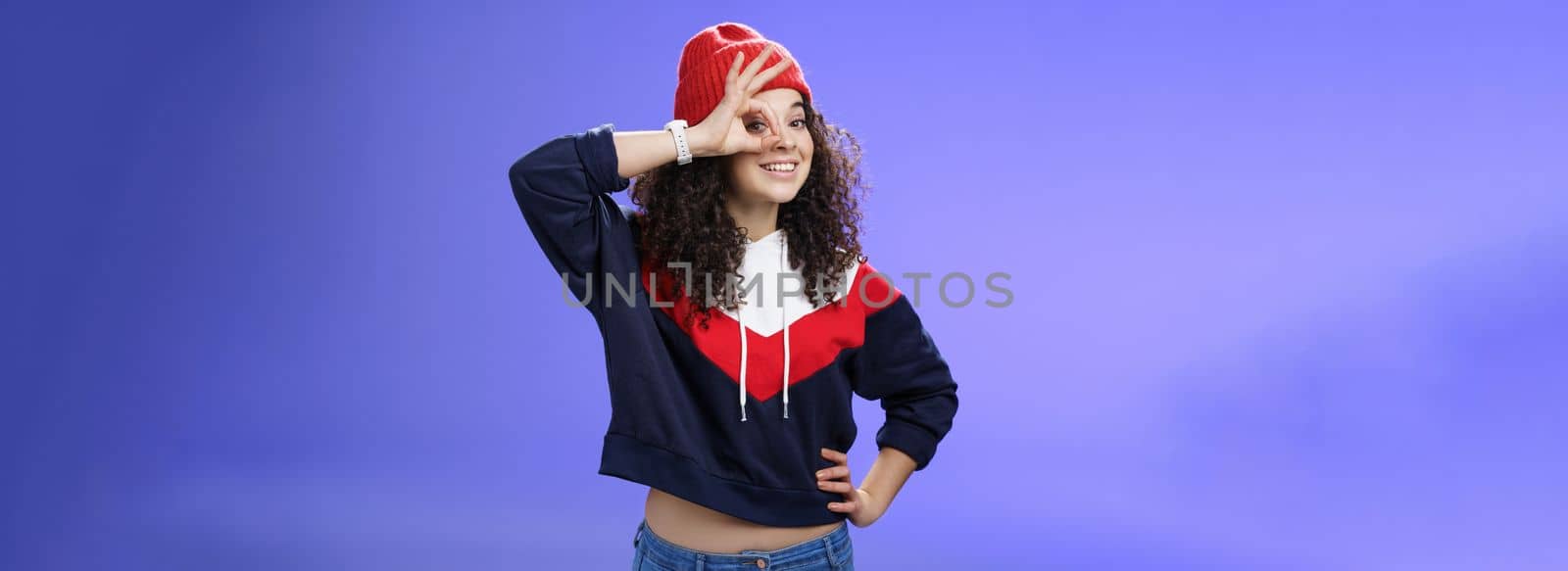 Okay girl agrees. Friendly-looking happy and charming young curly-haired girl in warm beanie showing ok gesture or circle over eye as peeking at camera and smiling broadly with joy over blue wall.