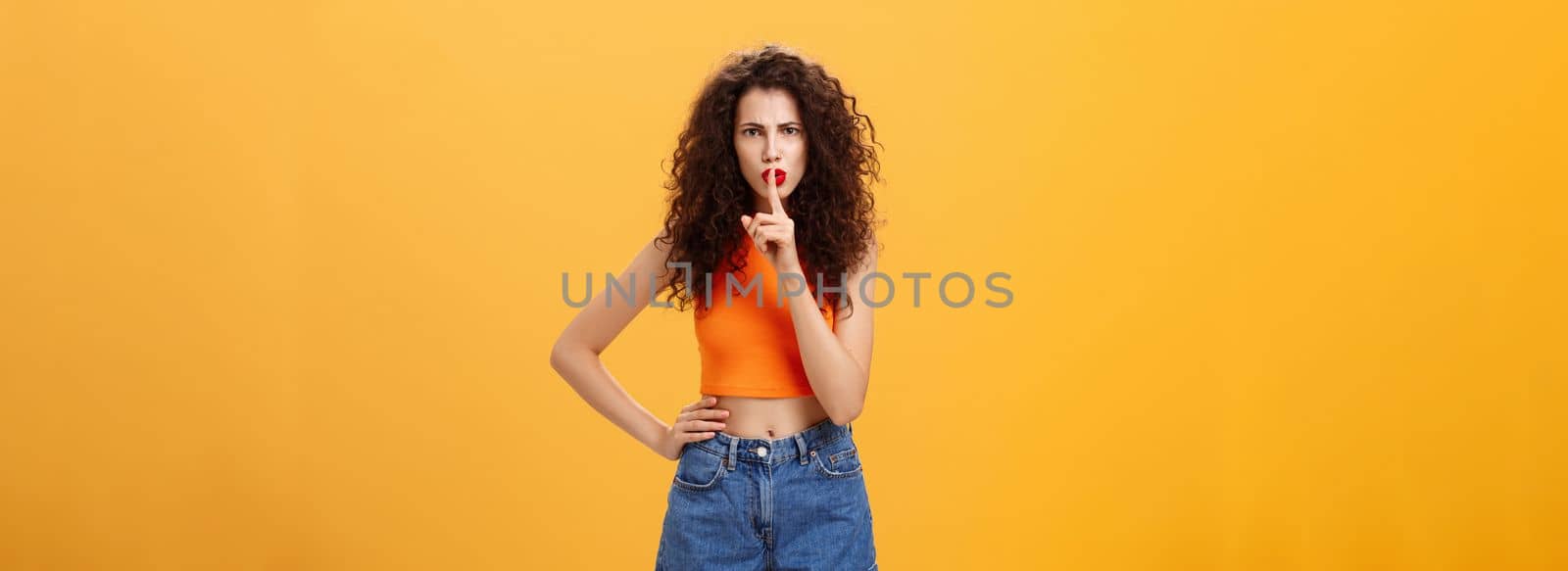 Sister displeased sibling saying cursing words standing serious and strict over orange background in stylish outfit shushing at camera saying shh with index finger over mouth demanding obey rules. Emotions concept
