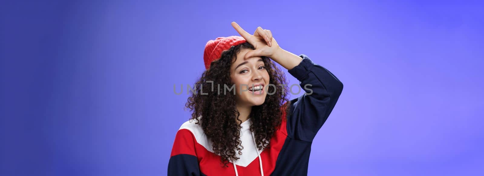 Girl triumphing and mocking friend as winning showing loser gesture on forehead and laughing having fun standing pleased and happy over blue background, smiling at camera joyfully by Benzoix