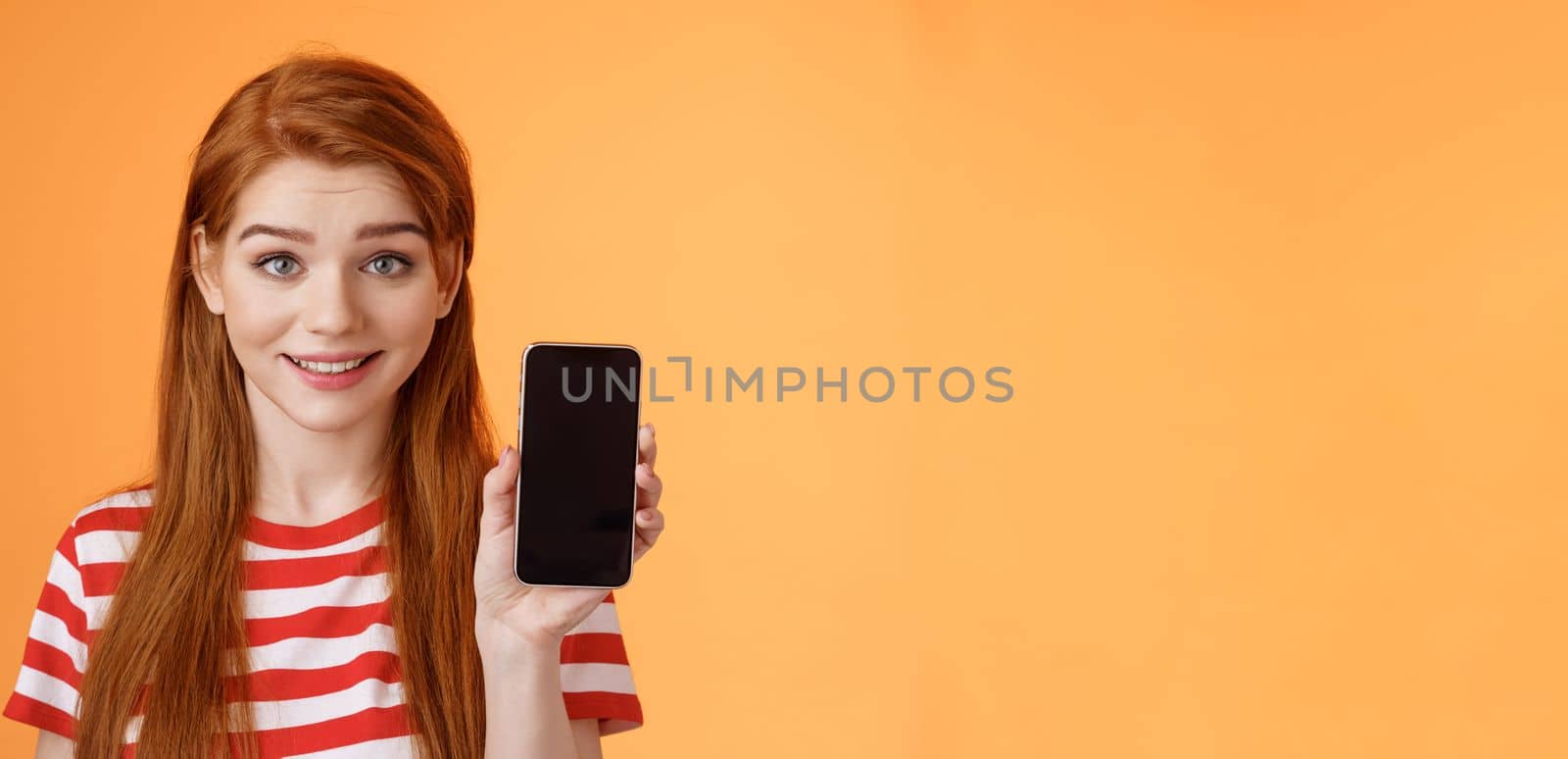 Close-up lovely caucasian redhead girl showing smartphone screen orange background, smiling cute, recommend application, promote app, online offer sale, introduce mobile phone. Technology concept